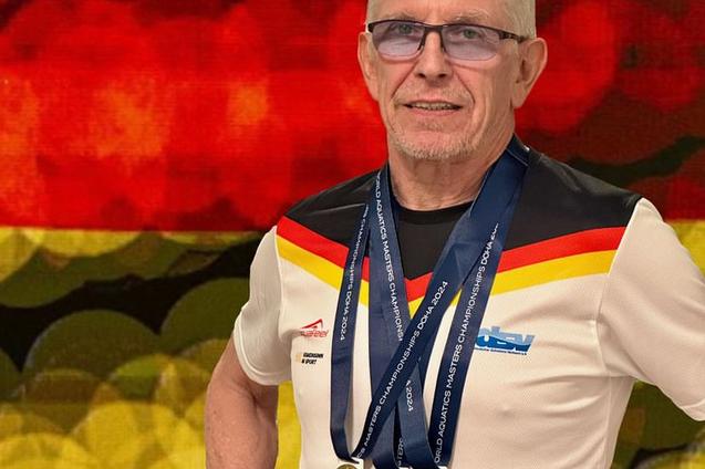 Mann mit grauem Haar tr&auml;gt ein sportliches Shirt und zeigt Medaillen auf rotem, unscharfem Hintergrund.