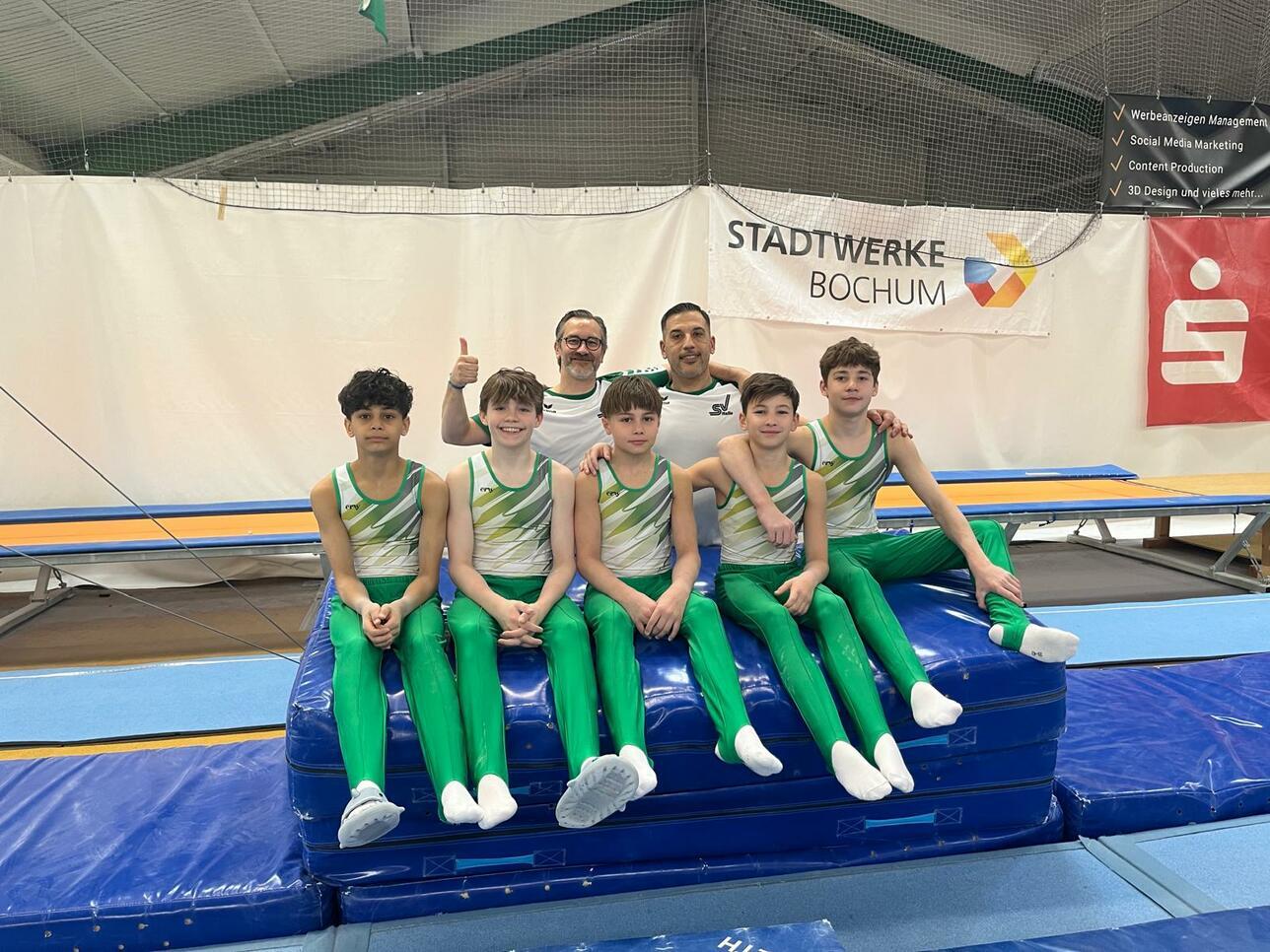 Gruppe von sechs Jungen in grünen Gymnastikanzügen auf einem blauen Kästchen, begleitet von einem Trainer im Hintergrund.