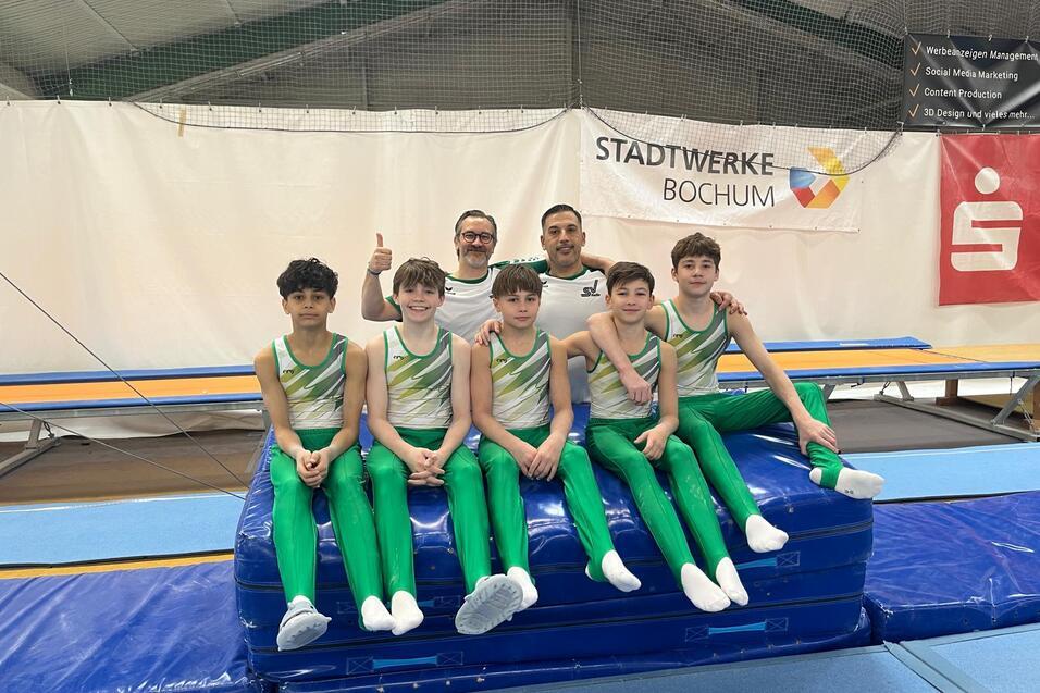 Gruppe von sechs Jungen in gr&uuml;nen Gymnastikanz&uuml;gen auf einem blauen K&auml;stchen, begleitet von einem Trainer im Hintergrund.