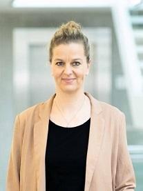 Portr&auml;t einer l&auml;chelnden Frau mit blonden Haaren, in einem beigen Blazer, vor einem modernen Hintergrund.