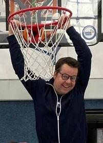 Mann hängt mit beiden Händen im Basketballkorb und lächelt, während er auf einem Sportplatz steht.