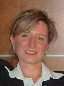 Portrait einer Frau mit blonden, leicht gewellten Haaren, die lächelt und einen hellen Kragen unter einem dunklen Oberteil trägt.