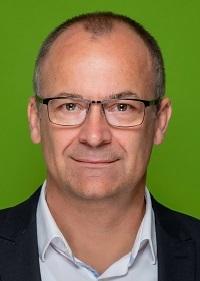 Porträt eines Mannes mit Brille vor einem grünen Hintergrund, lächelnd und im Anzug gekleidet.