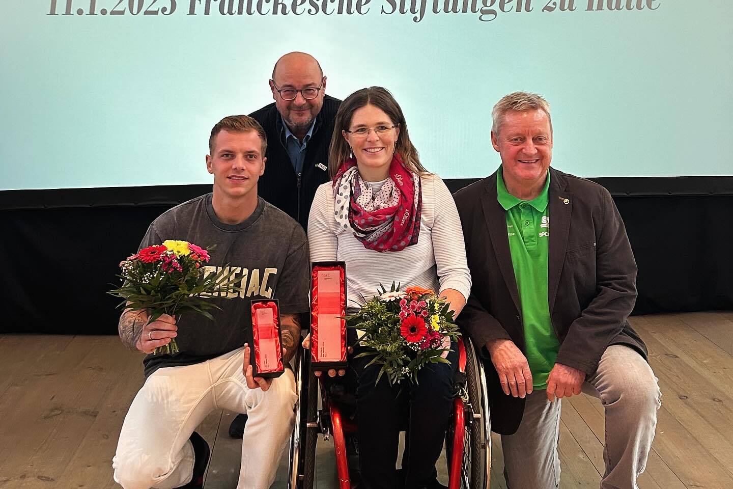 Vier Personen posieren mit Blumenstr&auml;u&szlig;en und Auszeichnungen vor einem Pr&auml;sentationshintergrund bei einer Sportveranstaltung.