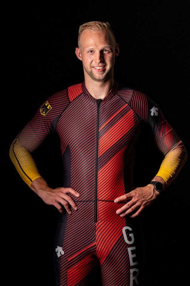 Lächelnder Athlet in einem rot-schwarzen Anzug mit deutschem Emblem, posierend mit Händen auf den Hüften vor schwarzem Hintergrund.