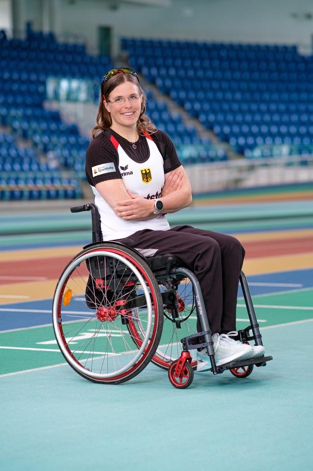 Porträt einer Sportlerin im Rollstuhl mit gekreuzten Armen auf einer bunten Laufbahn in einer Sportarena.