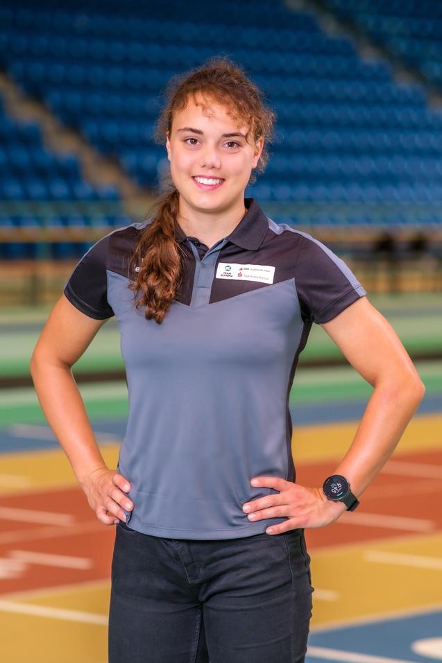 Porträt einer jungen Frau in Sportkleidung mit einer Armbanduhr, vor einem mehrfarbigen Sportboden.