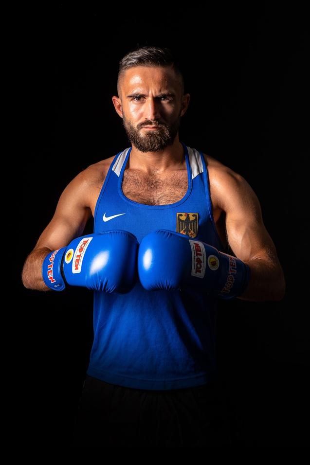 Ein Boxer in blauen Handschuhen und Trikot mit deutschem Wappen steht im Dunkeln und blickt ernst in die Kamera.