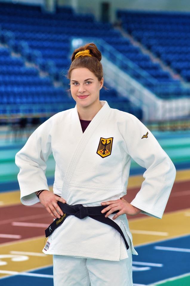 Junge Frau in Judogi mit schwarzem Gürtel steht auf einer Wettkampfmatte und lächelt. Hintergrund zeigt leere Tribünen.