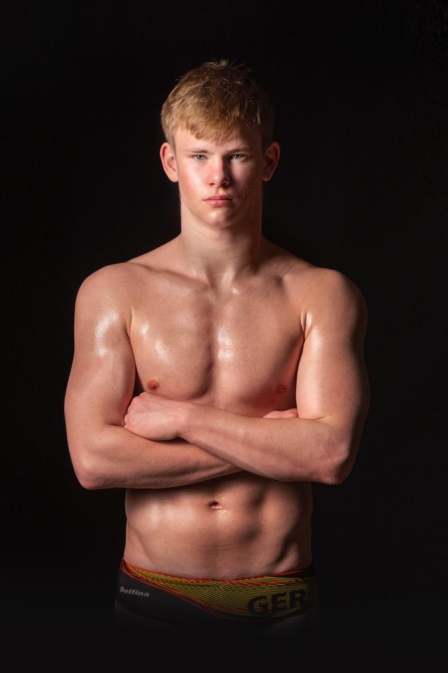 Junger, athletischer Mann mit muskulösem Oberkörper steht mit verschränkten Armen vor schwarzem Hintergrund.