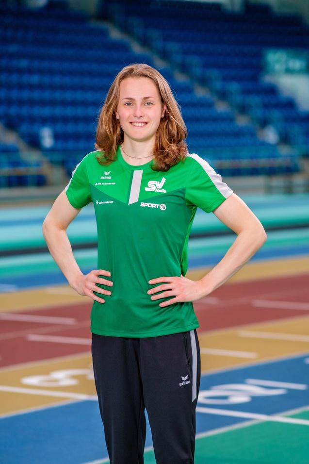 Sportlerin lächelt in grüner Trainingsbekleidung auf einer Laufbahn, Hände in die Hüften gestützt, Sporthalle im Hintergrund.