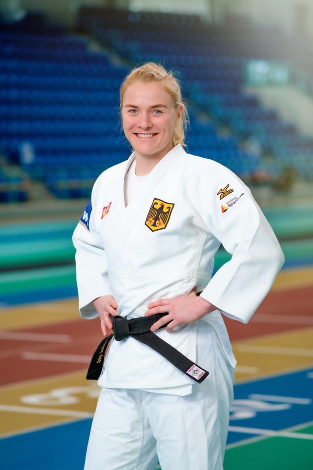 Junge Athletin in Judo-Uniform mit schwarzem Gürtel posiert lächelnd auf einer Judo-Trainingsmatte.