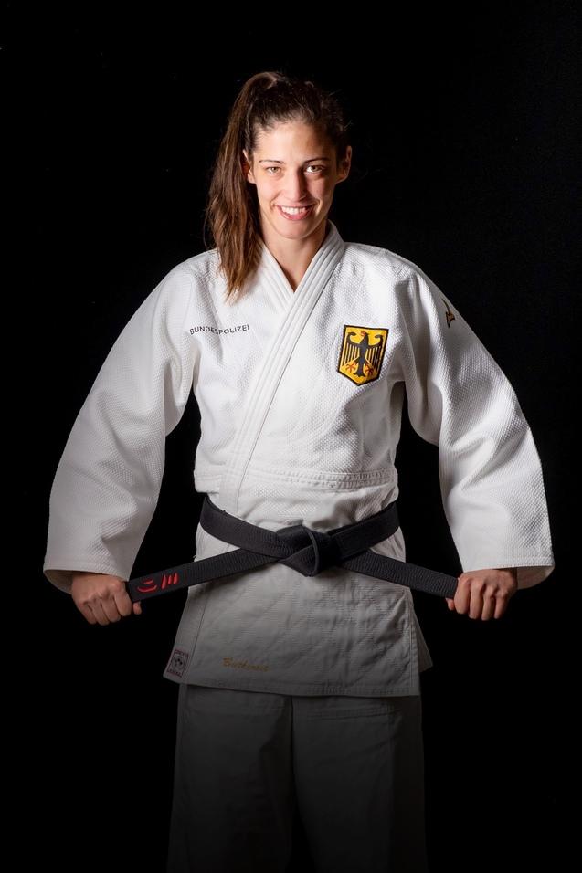 Eine lächelnde Judo-Athletin in einem weißen Gi mit schwarzem Gürtel posiert vor einem schwarzen Hintergrund.