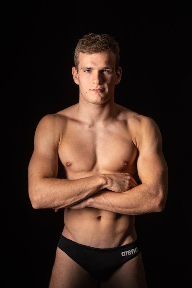 Junger Schwimmer mit durchtrainiertem Oberkörper, stehend mit verschränkten Armen vor schwarzem Hintergrund.