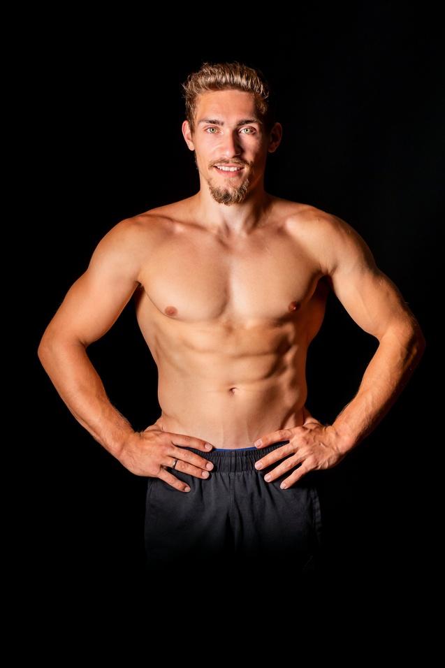 Mann mit muskulösem Oberkörper steht mit Händen auf den Hüften vor schwarzem Hintergrund und lächelt.