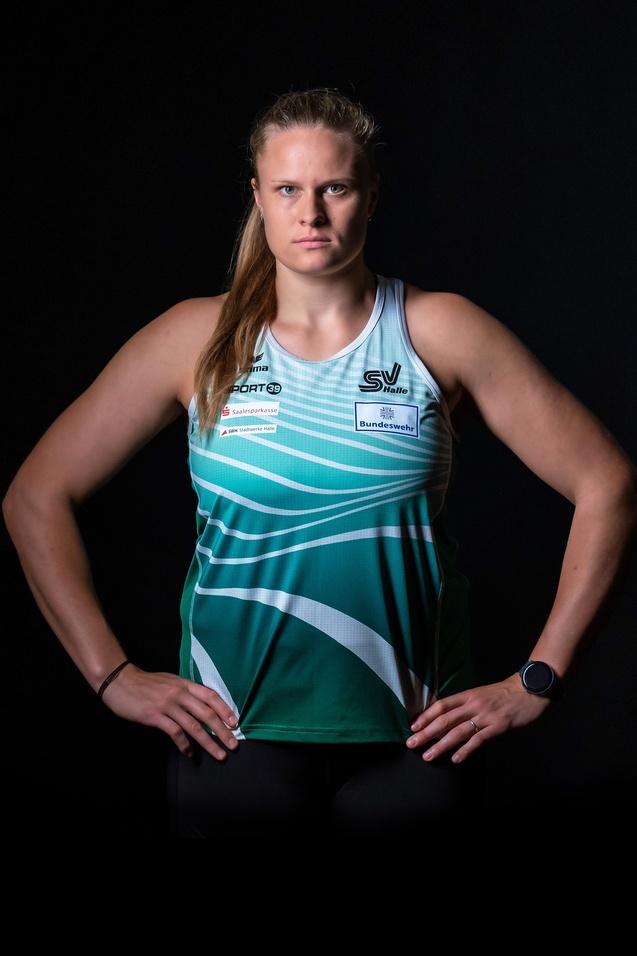 Frau mit resolutem Gesichtsausdruck trägt ein farbiges Sporttrikot und steht mit verschränkten Armen vor schwarzem Hintergrund.