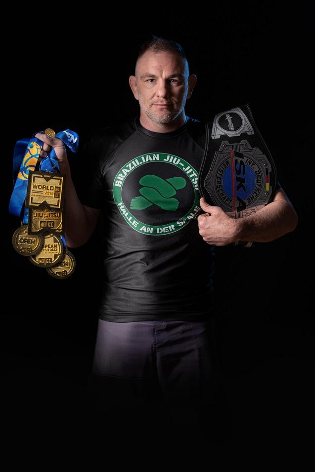 Mann im schwarzen T-Shirt mit "Brazilian Jiu-Jitsu"-Logo, hält Medaillen und einen Gürtel in der Hand.
