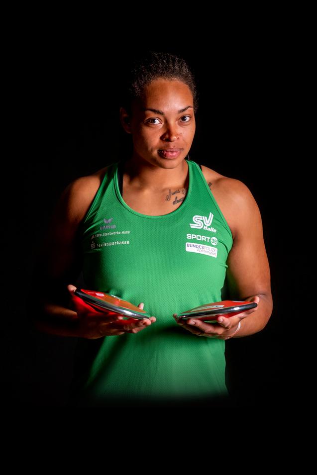 Frau in grünem Sporttrikot hält zwei Tischtennisschläger mit Bällen in der Hand, vor einem schwarzen Hintergrund.