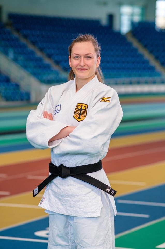 Junge Frau im Judogi mit schwarzem Gürtel steht mit verschränkten Armen auf einer Sportfläche.