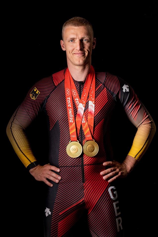 Athlet in einem rot-schwarzen Anzug präsentiert stolz zwei olympische Goldmedaillen vor schwarzem Hintergrund.