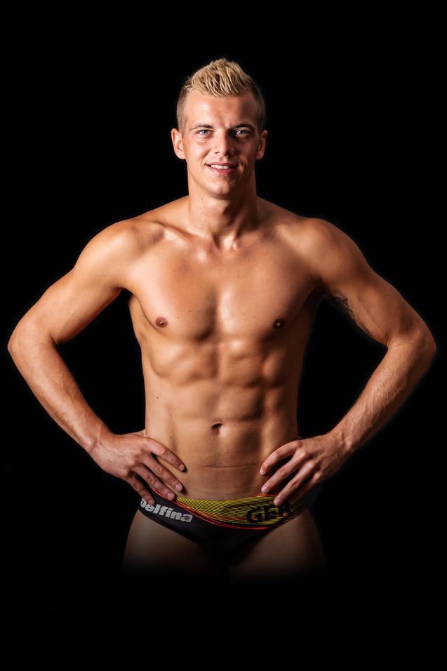 Junger, muskulöser Mann steht mit freiem Oberkörper und Händen an der Hüfte vor schwarzem Hintergrund.