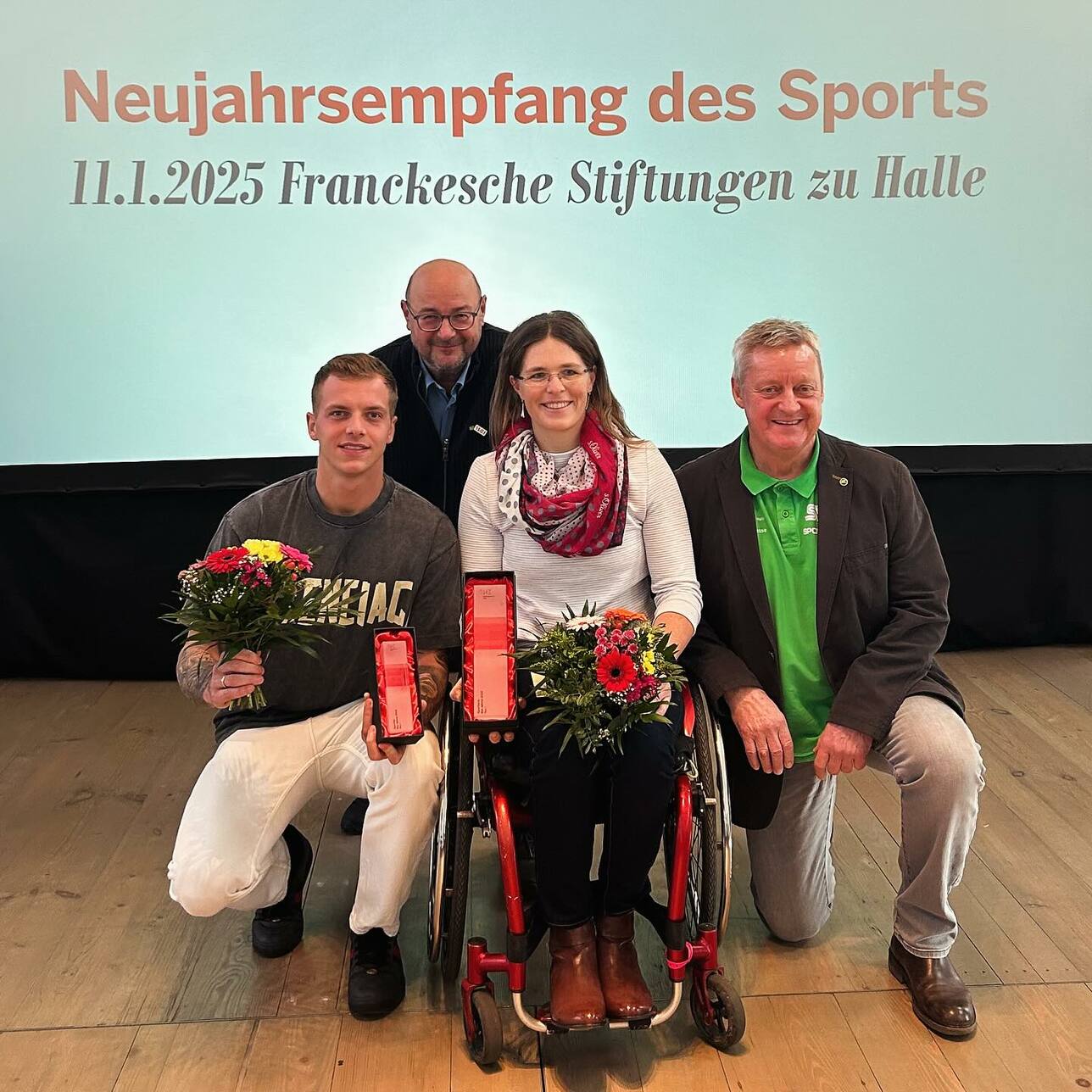 Vier Personen posieren mit Blumensträußen und Auszeichnungen vor einem Präsentationshintergrund bei einer Sportveranstaltung.