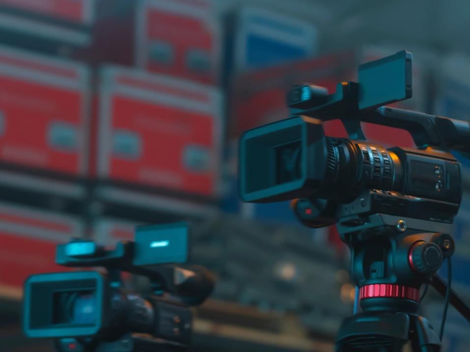 Drei Videokameras auf Stativen in einem Lager mit bunten Kisten im Hintergrund.