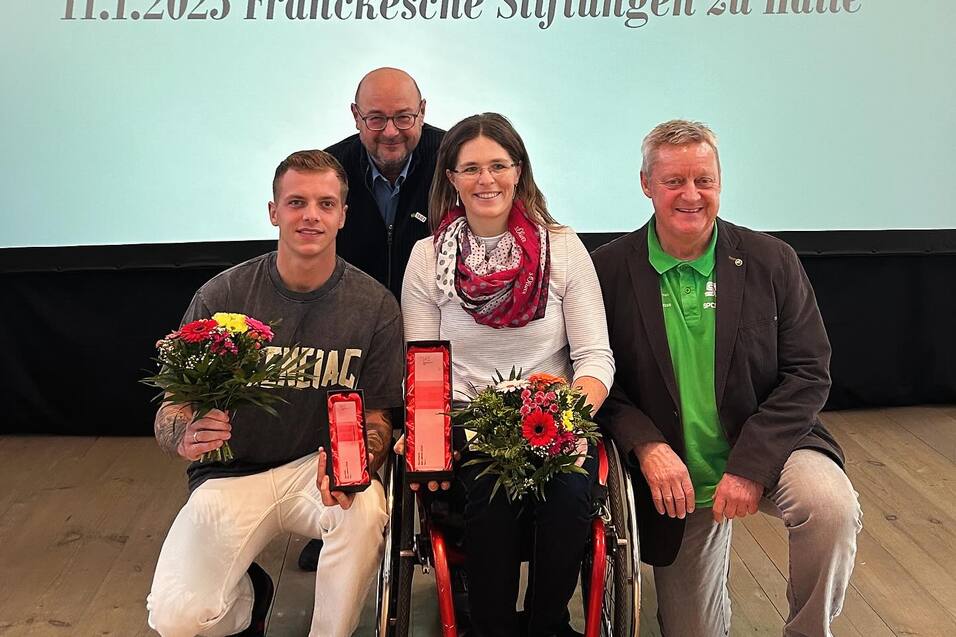 Vier Personen posieren mit Blumensträußen und Auszeichnungen vor einem Präsentationshintergrund bei einer Sportveranstaltung.