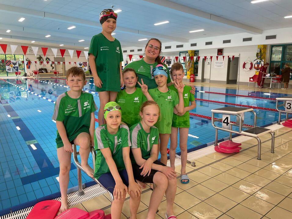 Gruppenselfie von sieben Kindern und einer Trainerin am Schwimmbecken, alle tragen gr&uuml;ne Schwimmanz&uuml;ge.