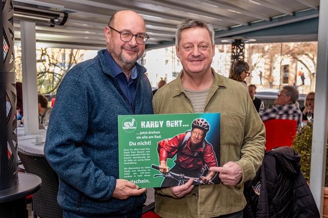 Zwei M&auml;nner stehen in einem Restaurant und halten ein Plakat mit einem Radfahrer und einem Werbeslogan.