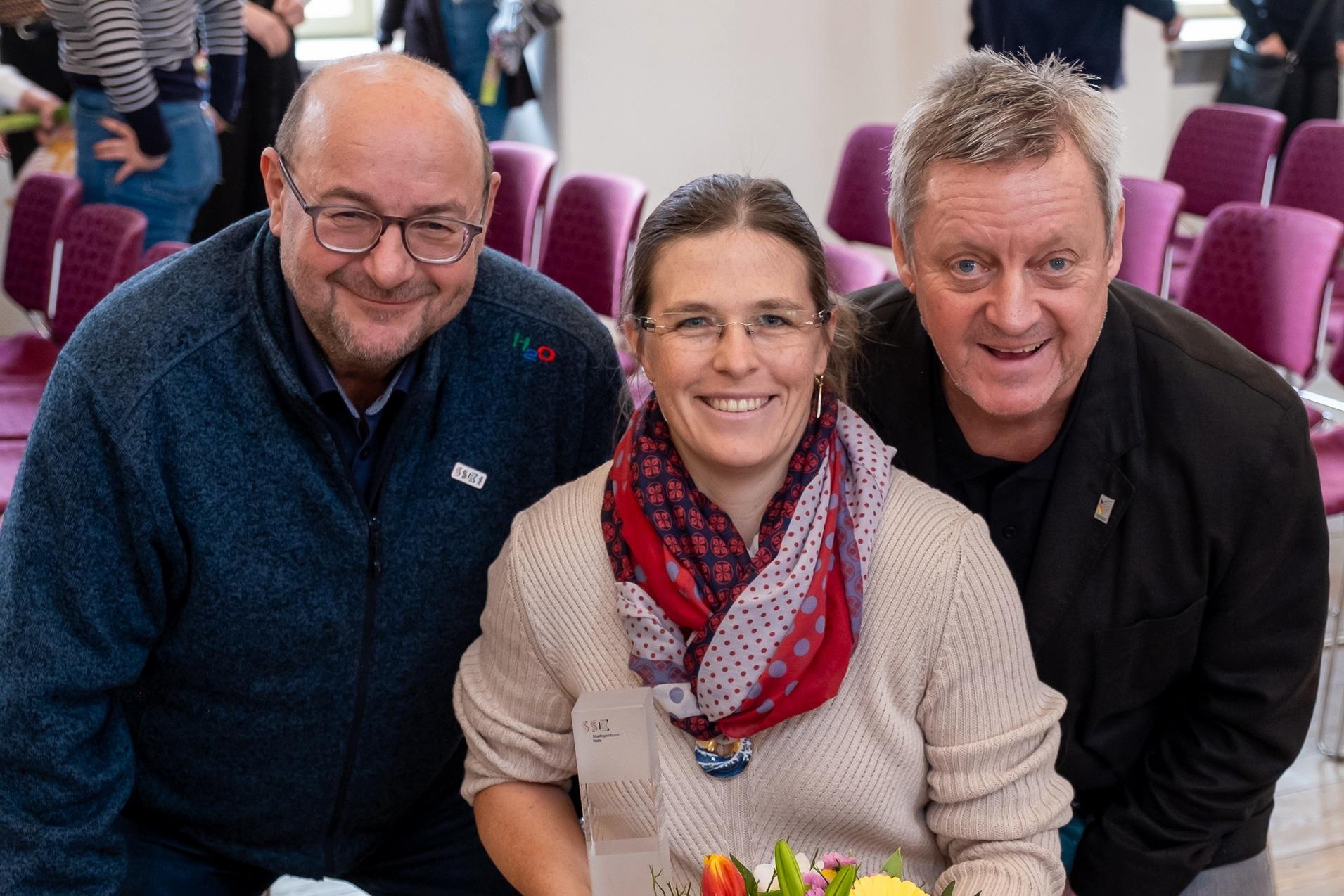 Drei Personen posieren in einem Raum, eine Frau mit Blumen und einem Preis, von zwei Männern flankiert.
