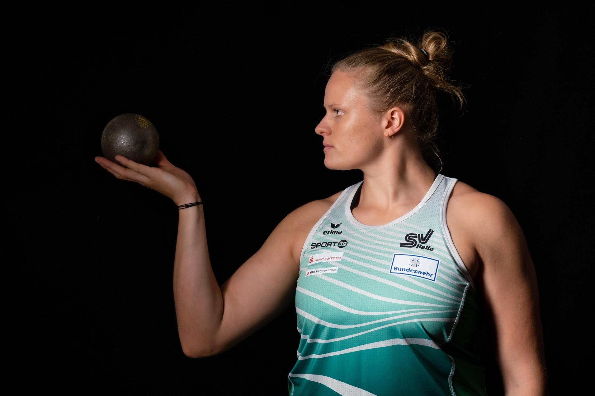 Athletin h&auml;lt eine Kugel in der Hand, schaut konzentriert zur Seite, schwarze Hintergrundbeleuchtung.