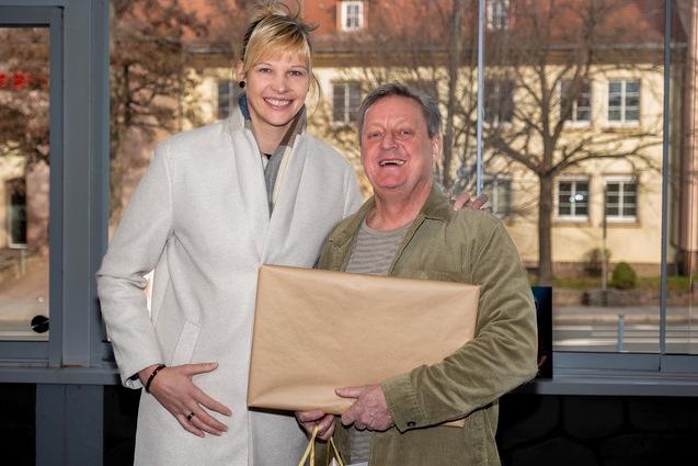 Ein Mann und eine Frau lächeln vor einem Fenster, der Mann hält ein Geschenk in braunem Papier.
