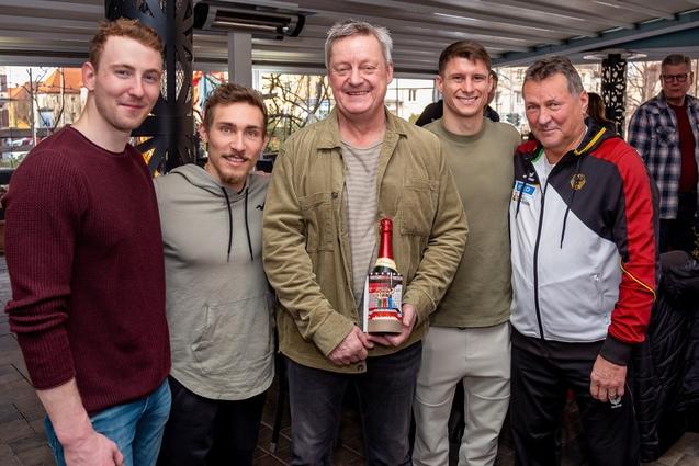 Gruppenfoto von fünf Männern, einer hält eine Flasche Sekt, umgeben von lächelnden Freunden in lockerer Umgebung.