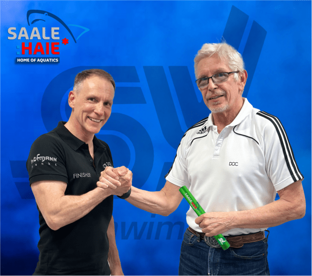 Zwei Männer schütteln sich die Hände vor einem blauen Hintergrund mit dem Logo „Saale Haie“. Der zweite Mann hält einen grünen Gegenstand.
