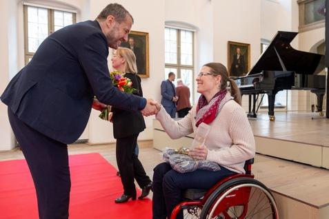 Eine Frau im Rollstuhl sch&uuml;ttelt die Hand eines Mannes, der einen Blumenstrau&szlig; h&auml;lt, auf einem roten Teppich.