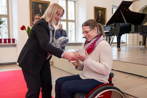 Frau im Rollstuhl erh&auml;lt von einer Dame ein Geschenk in einem festlichen Raum mit Klavier und Bildern an der Wand.