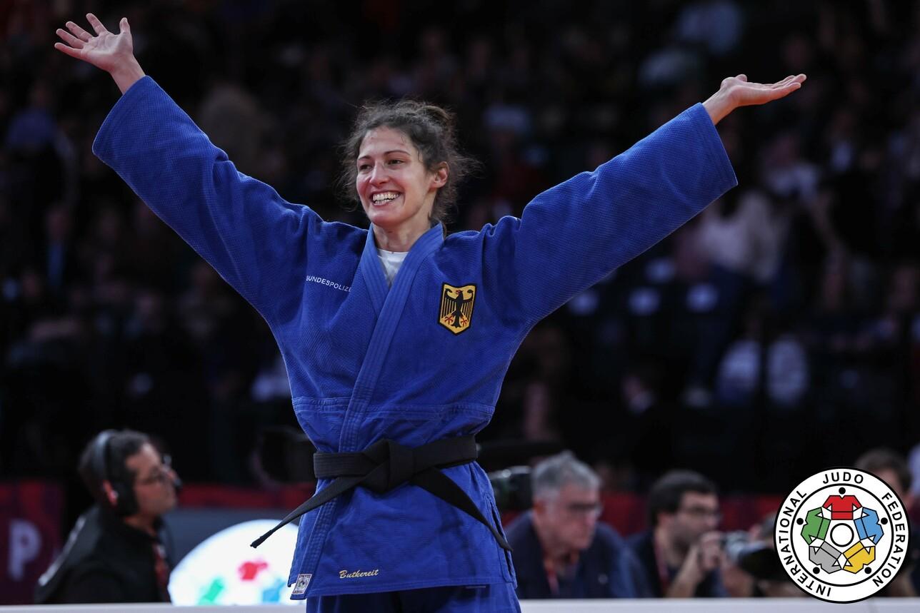 Judo-Athletin in blauer Gi-Kleidung hebt beide Arme freudig in die Luft, während sie in einer Wettkampfarena steht.