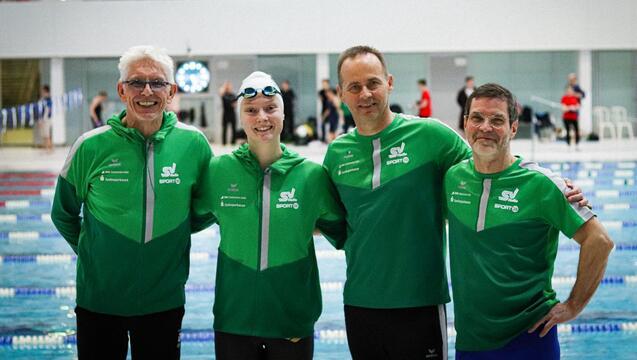 Vier Schwimmer in gr&uuml;nen Sportjacken stehen am Beckenrand eines Schwimmbades und l&auml;cheln in die Kamera.