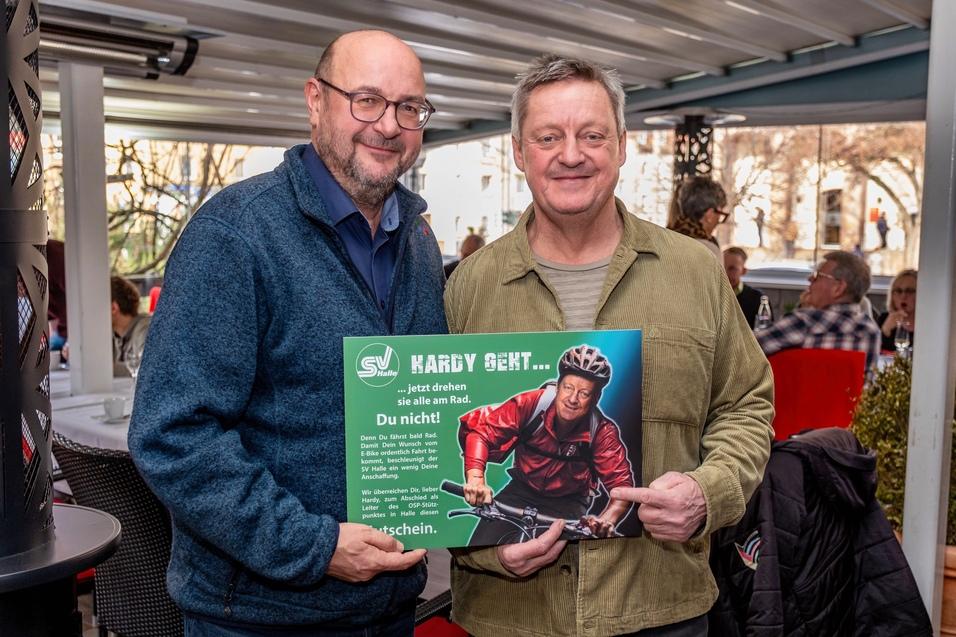 Zwei M&auml;nner stehen in einem Restaurant und halten ein Plakat mit einem Radfahrer und einem Werbeslogan.