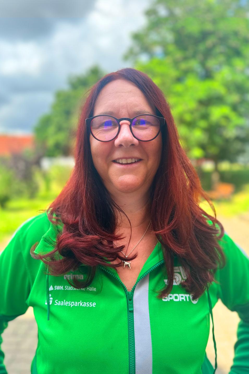 Frau mit langen, roten Haaren und Brille, in gr&uuml;ner Sportjacke, vor verschwommenem, pflanzenreichen Hintergrund.