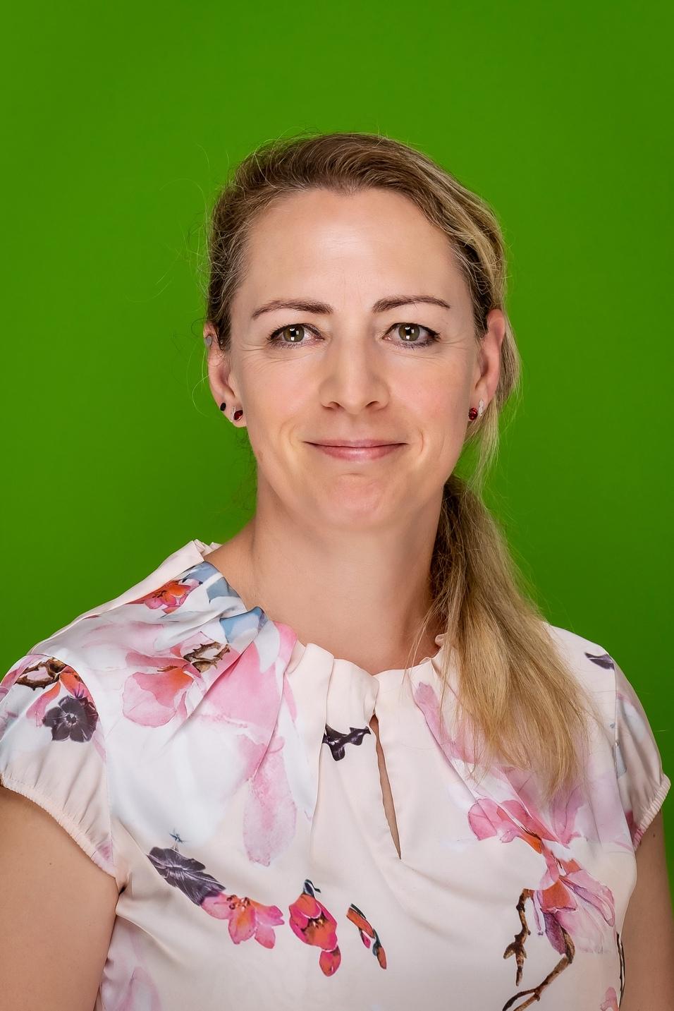 Portr&auml;t einer Frau mit blonden Haaren, die ein blumiges Oberteil tr&auml;gt, vor einem einfarbigen gr&uuml;nen Hintergrund.