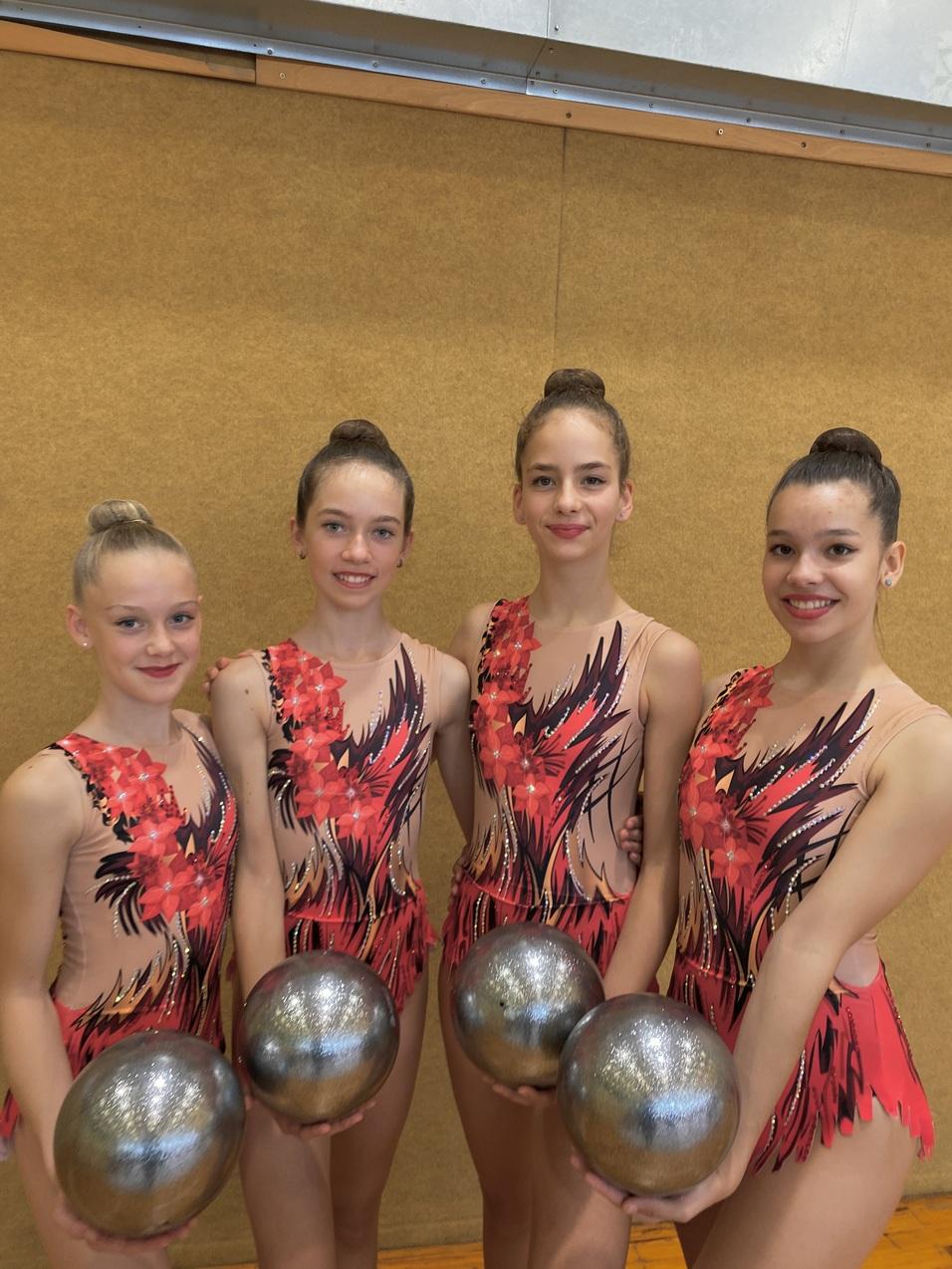 Vier junge rhythmische Sportlerinnen in bunten Kostümen halten silberne Bälle vor einem hellbraunen Hintergrund.