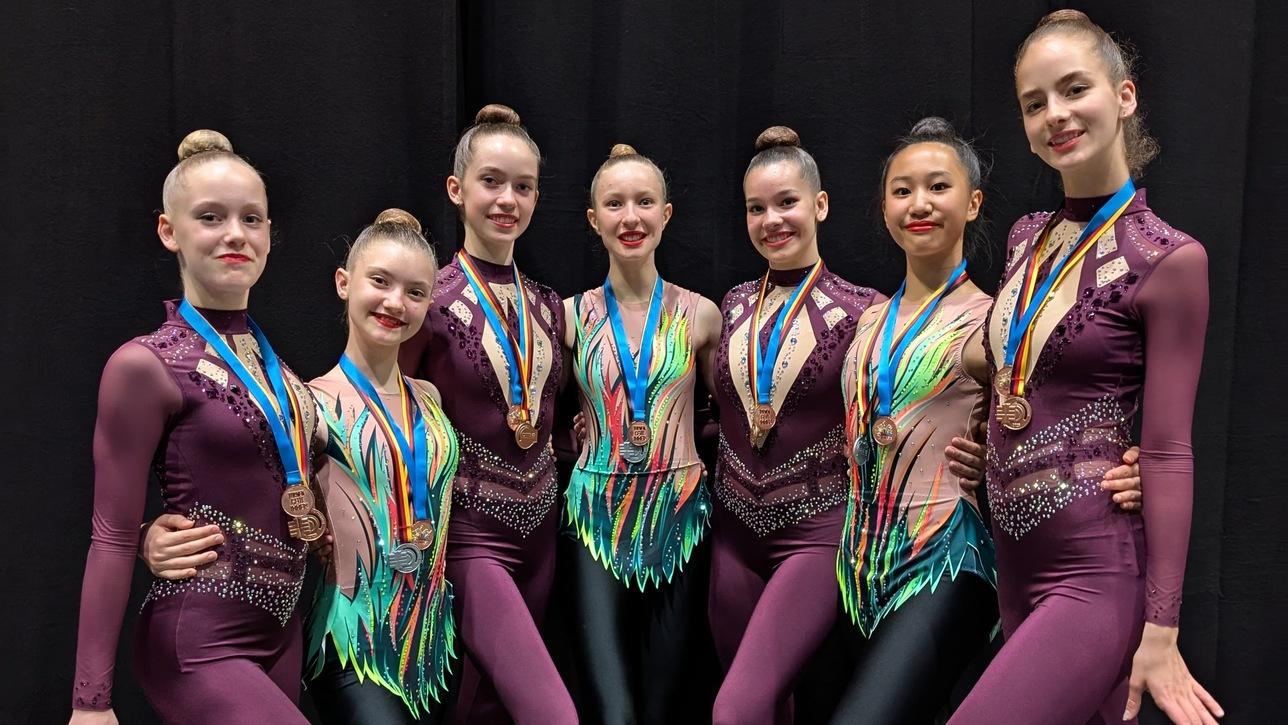Sieben Tänzerinnen in bunten Kostümen mit Medaillen posieren fröhlich vor schwarzem Hintergrund.