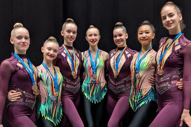 Sieben T&auml;nzerinnen in bunten Kost&uuml;men mit Medaillen posieren fr&ouml;hlich vor schwarzem Hintergrund.