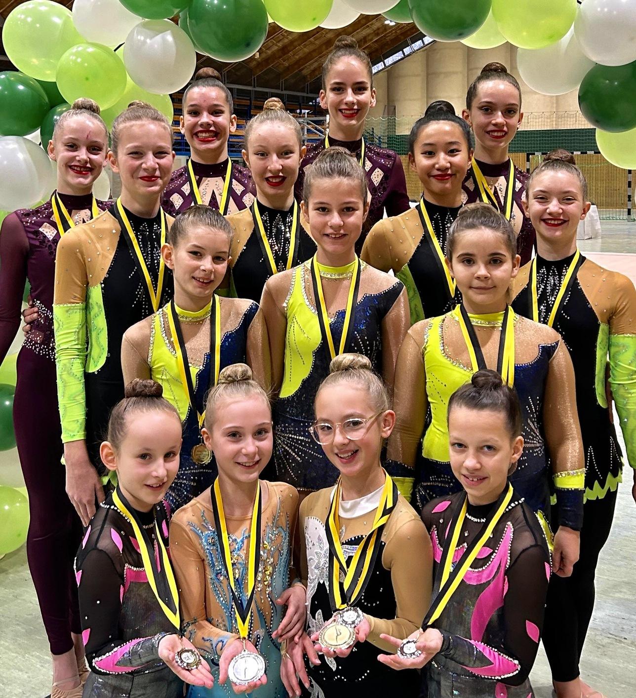 Gruppe von Tänzerinnen in bunten Kostümen mit Medaillen, stehend vor einem dekorativen Luftballonbogen.