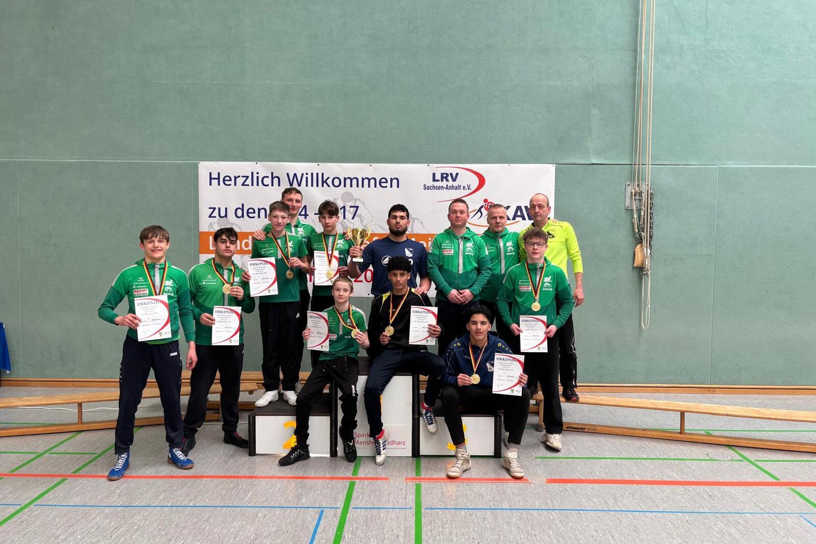 Sieben Medaillengewinner in gr&uuml;nen Sportjacken halten Urkunden auf einem Podest vor einem Willkommensbanner.