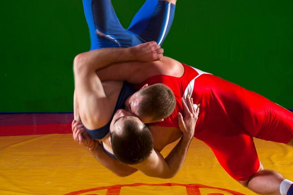Zwei Ringer kämpfen auf einer gelben Matte, einer im blauen, der andere im roten Anzug, mit grünem Hintergrund.