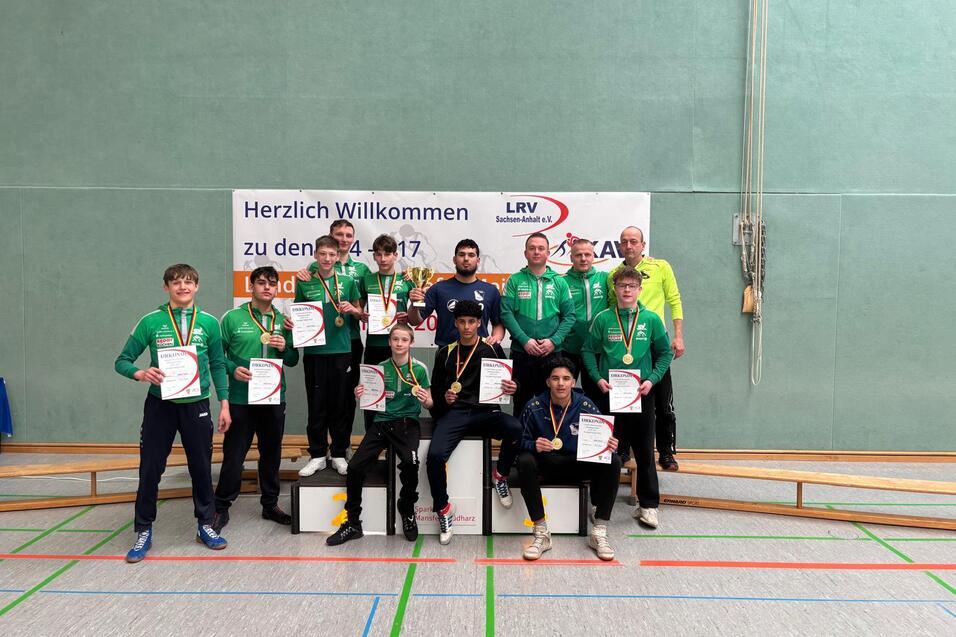 Sieben Medaillengewinner in gr&uuml;nen Sportjacken halten Urkunden auf einem Podest vor einem Willkommensbanner.