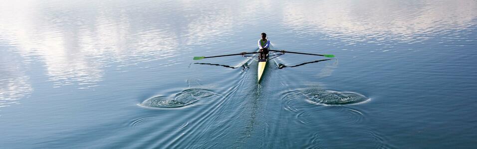 Ruderer in einem Einzelboot paddelt über einen ruhigen See, Wasseroberfläche spiegelt den Himmel wider.
